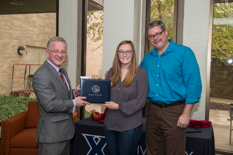 Library Director Ken Gibson, Sydney Sanders, and Dr. Graley Herren smiling together in the Xavier University Library. Gibson and Sanders are holding the award together.